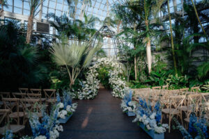 Lush indoor garden wedding ceremony with a floral aisle, tropical greenery, and soft blue floral accents.