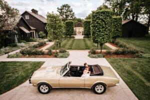 Couple arriving at a garden wedding venue in a vintage convertible, framed by manicured grounds and classic architecture.