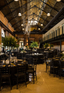 Industrial wedding reception space with long tables, black chairs, greenery installations, and dramatic overhead lighting.